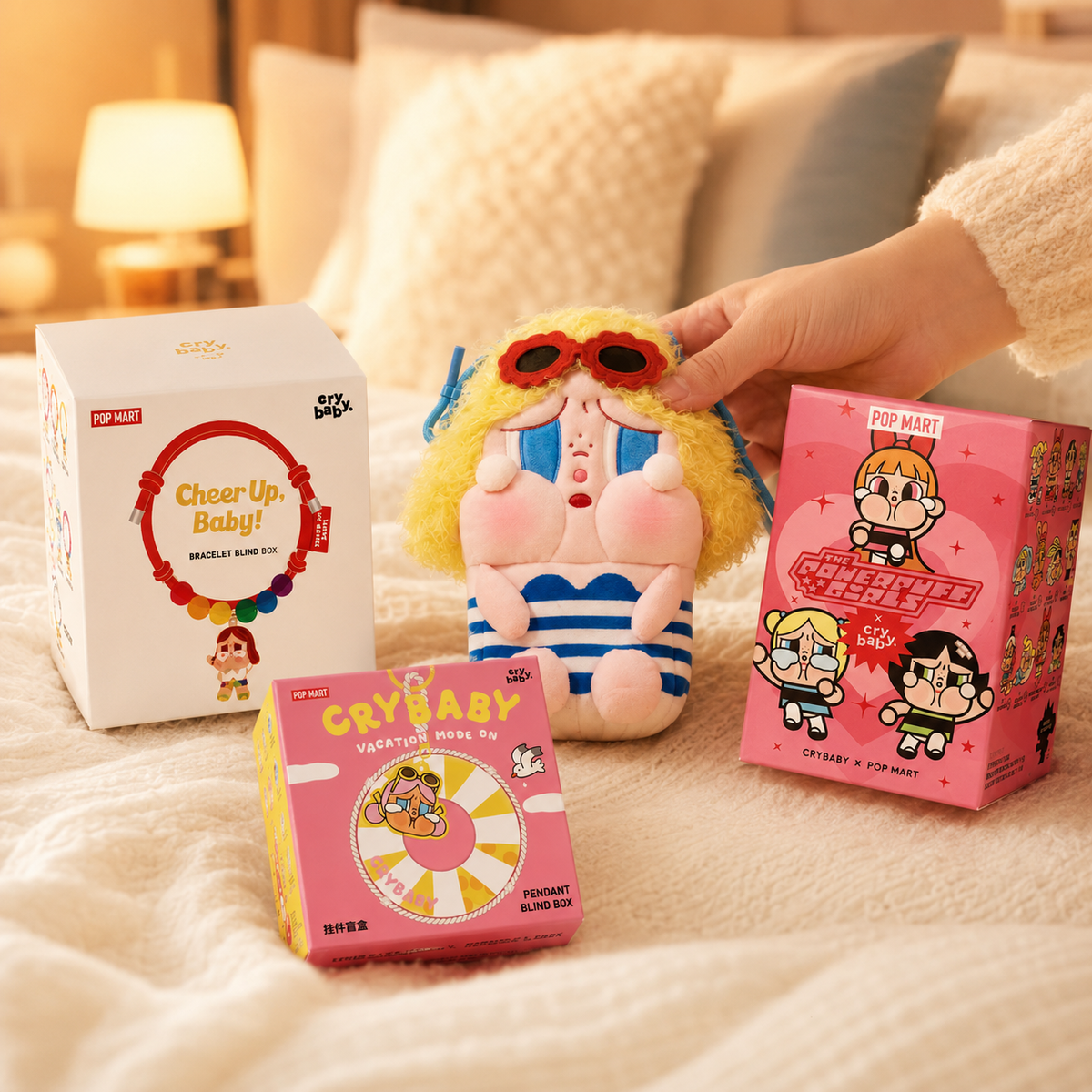 Hand holding a POP MART CRYBABY Soft Hearts plush toy beside two blind boxes and a pink accessory box on a bed.
